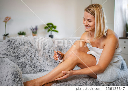 Smiling woman shaving her legs with an electric razor sitting on the sofa Smiling woman shaving her legs with an electric razor sitting on the sofa 136127847