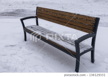 an icy, snow-covered park empty bench with icicles hanging down 136129331