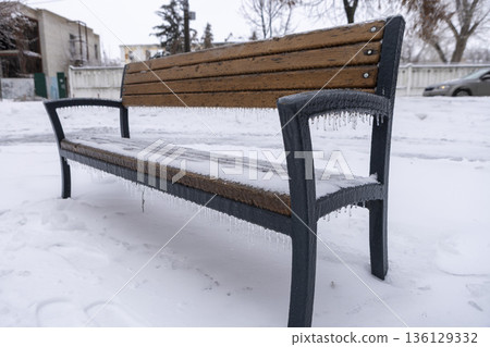 an icy, snow-covered park empty bench with icicles hanging down 136129332