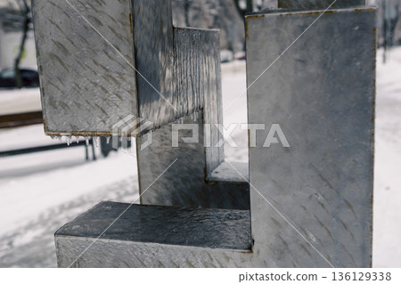 rusted metal frame overlooking snowy sidewalk, shallow depth emphasizes blurred urban path beyond, flaked paint and ice crystals clinging to edge, muted winter 136129338