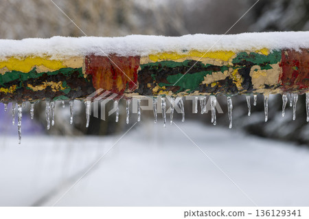 Close-up of a frost-covered multicolored metal railing with small hanging icicles in winter. Close-up of a frost-covered multicolored metal railing with small hanging icicles in winter. 136129341