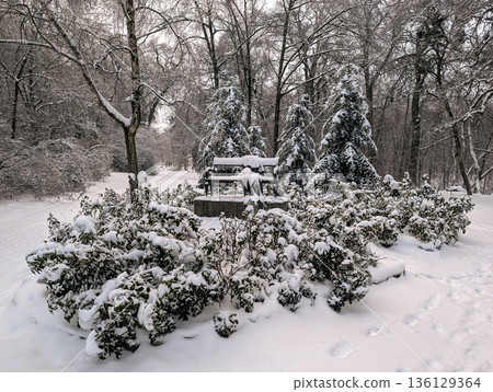 In a snowy winter park, a sculpture shaped like a bench with clothes stands beneath falling snow. 136129364