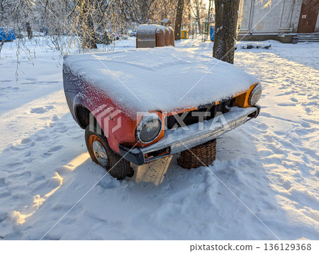 A snow covered vintage car abandoned in front, orange faded paint and rusty grille, missing headlights, one front wheel lying in the packed snow, an old chain hanging. A snow covered vintage car abandoned in front, orange faded paint and rusty grille, missing headlights, one front wheel lying in the packed snow, an old chain hanging. 136129368