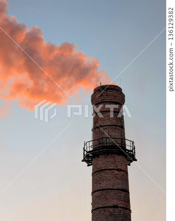 a heating plant chimney with a column of smoke pouring out, illuminated by the orange setting sun a heating plant chimney with a column of smoke pouring out, illuminated by the orange setting sun 136129382