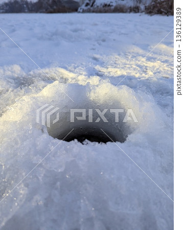 An ice-covered river with a fresh fishing hole under the cold winter sun. 136129389