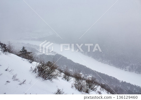 Snowy mountain slope overlooking a frozen river and distant village. Winter landscape under heavy snowfall for nature and travel. 136129460