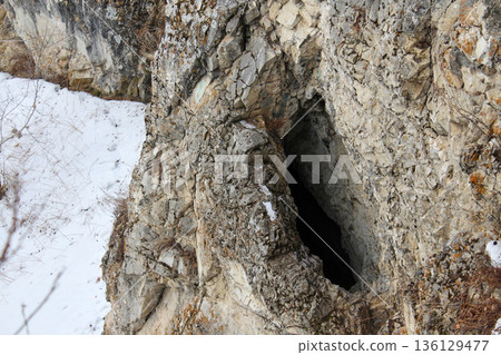 Natural cave entrance in a rocky cliff wall during winter with snow. Geological formation concept for nature exploration. Natural cave entrance in a rocky cliff wall during winter with snow. Geological formation concept for nature exploration. 136129477