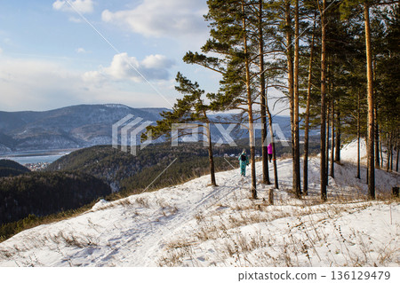 Man and woman hiking on a snowy trail in the siberian taiga. Winter forest with pine trees and mountains for winter travel. Man and woman hiking on a snowy trail in the siberian taiga. Winter forest with pine trees and mountains for winter travel. 136129479