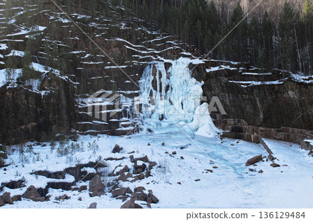 Frozen waterfall on a cliff partially covered with snow and green trees. Winter landscape in Krasnoyarsk Stolby national park, Russia. 136129484