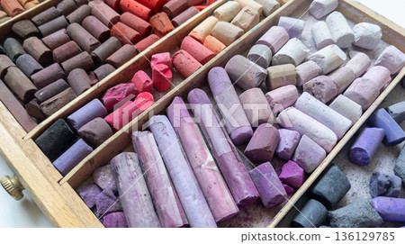 Broken soft pastel crayons in a wooden box, top view. Lilac and red shades arranged by tone. Artist's art supplies. Broken soft pastel crayons in a wooden box, top view. Lilac and red shades arranged by tone. Artist's art supplies. 136129785