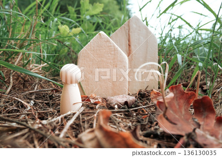 Wooden figure standing near two wooden house shapes in a natural outdoor setting Wooden figure standing near two wooden house shapes in a natural outdoor setting 136130035