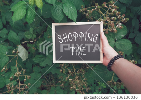 Hand holding chalkboard sign with text shopping time surrounded by lush green foliage Hand holding chalkboard sign with text shopping time surrounded by lush green foliage 136130036