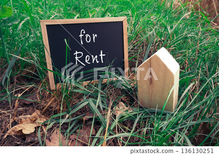 Wooden house model beside chalkboard sign indicating property for rent in grassy area 136130251