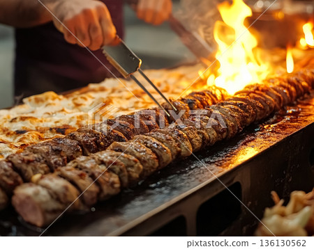 Grilled meat skewers sizzling on a barbecue with flames and delicious food in the background 136130562