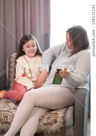 A mother and daughter share a joyful moment, looking at a phone together on a comfy couch. 136131181