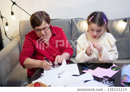 Grandmother and granddaughter create paper crafts together, enjoying a cozy moment of shared creativity. Grandmother and granddaughter create paper crafts together, enjoying a cozy moment of shared creativity. 136131214