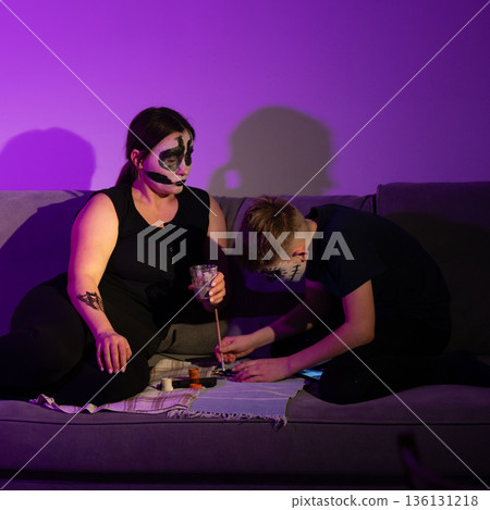A woman and child, adorned in spooky face paint, relax on a couch, engaging in a creative activity together. 136131218
