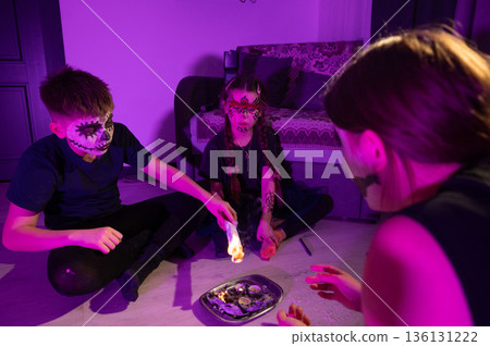 Kids with spooky face paint gather around a burning candle during a Halloween celebration. 136131222