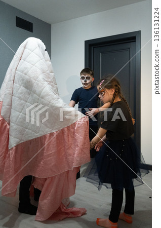Children playfully interact with a person hidden under a sheet, creating a ghostly Halloween scene. 136131224
