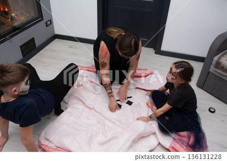A parent and two children with Halloween face paint arrange black shapes on a blanket, seemingly creating a game or decoration. A parent and two children with Halloween face paint arrange black shapes on a blanket, seemingly creating a game or decoration. 136131228