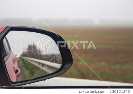 Rear view mirror in car. Car on highway on cloudy day. Car mirror reflection in the field. Auto travel concept. Car trip concept. Part of vehicle. Rear view mirror in car. Car on highway on cloudy day. Car mirror reflection in the field. Auto travel concept. Car trip concept. Part of vehicle. 136131240