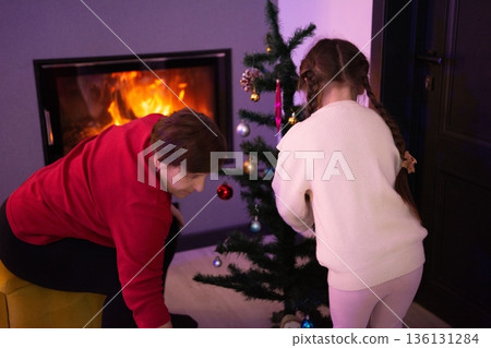 Grandmother and granddaughter decorate a small Christmas tree together by a fireplace, enjoying the cozy holiday atmosphere. Grandmother and granddaughter decorate a small Christmas tree together by a fireplace, enjoying the cozy holiday atmosphere. 136131284