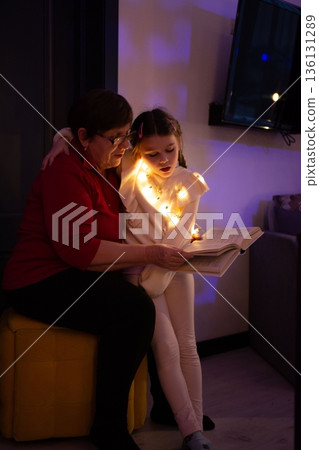 Grandmother and granddaughter snuggle together, illuminated by fairy lights, sharing a storybook. Grandmother and granddaughter snuggle together, illuminated by fairy lights, sharing a storybook. 136131289