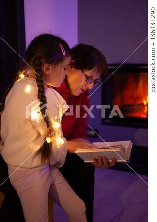 A grandmother and granddaughter snuggle by a fireplace, sharing a book and the warmth of the evening. A grandmother and granddaughter snuggle by a fireplace, sharing a book and the warmth of the evening. 136131290