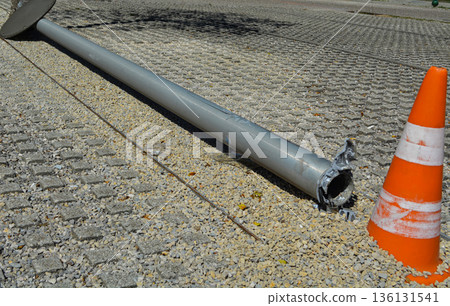 A broken streetlight pole on the ground in an urban area, marked by an orange traffic cone for safety. Concept: public safety, road hazards, city services, and urban restoration. A broken streetlight pole on the ground in an urban area, marked by an orange traffic cone for safety. Concept: public safety, road hazards, city services, and urban restoration. 136131541