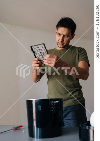 Vertical portrait of young man carefully inspecting and cleaning air humidifier filter, ensuring clean and healthy air in home environment, performing regular maintenance for optimal performance. Vertical portrait of young man carefully inspecting and cleaning air humidifier filter, ensuring clean and healthy air in home environment, performing regular maintenance for optimal performance. 136132860
