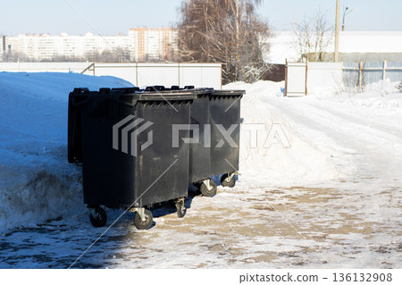 Municipal waste containers stand on frozen ground under winter sunlight near apartment complexes Municipal waste containers stand on frozen ground under winter sunlight near apartment complexes 136132908