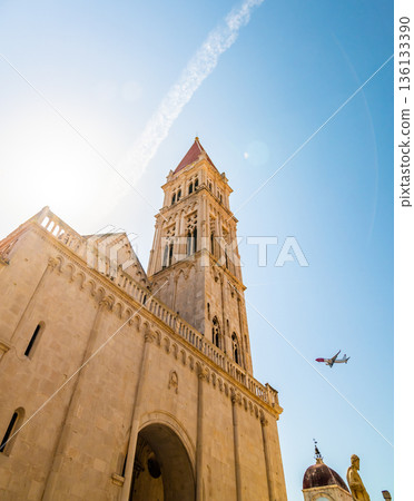 View of cathedral of St Lawrence, Trogir - Croatia. Down to up look to beautiful ancient church tower. The airplane is landing to Split airport. Aircraft is visible near the tower. View of cathedral of St Lawrence, Trogir - Croatia. Down to up look to beautiful ancient church tower. The airplane is landing to Split airport. Aircraft is visible near the tower. 136133390
