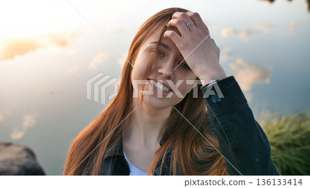 Smiling young woman playing with hair by lake at golden hour, expressing happiness Smiling young woman playing with hair by lake at golden hour, expressing happiness 136133414