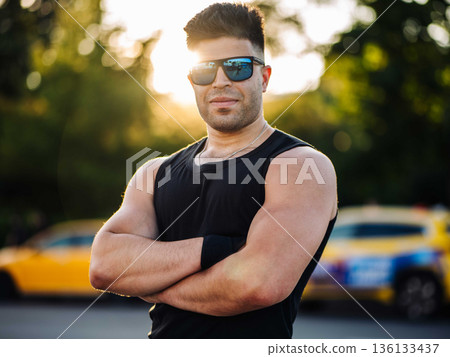Confident man with sunglasses and muscular arms standing outdoors at golden hour Confident man with sunglasses and muscular arms standing outdoors at golden hour 136133437