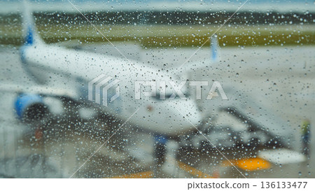 Raindrops blanketing airport window glass, blurring view of parked airplane, conveying travel anticipation and momentary stillness Raindrops blanketing airport window glass, blurring view of parked airplane, conveying travel anticipation and momentary stillness 136133477