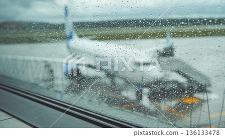Raindrops covering airport window blurring sight of parked airplane against gray, wet tarmac backdrop Raindrops covering airport window blurring sight of parked airplane against gray, wet tarmac backdrop 136133478