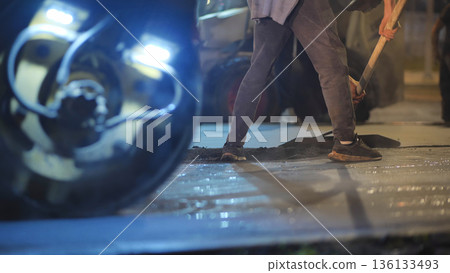 Road worker spreading hot asphalt at night with paving machine in city 136133493