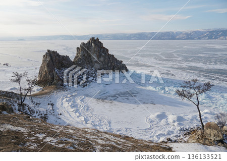 Wide winter view of Shaman Rock on Olkhon Island, Lake Baikal, surrounded by frozen lake surface, snow patterns, and distant mountains 136133521