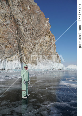Woman in warm winter clothing stands on clear ice near massive frozen cliffs of Lake Baikal under bright blue sky Woman in warm winter clothing stands on clear ice near massive frozen cliffs of Lake Baikal under bright blue sky 136133523