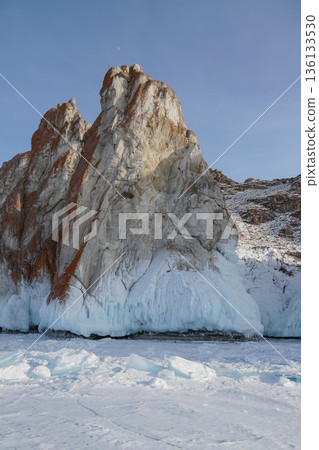 Massive rocky cliff rises from the frozen surface of Lake Baikal, covered with thick ice formations and snow under a clear blue winter sky Massive rocky cliff rises from the frozen surface of Lake Baikal, covered with thick ice formations and snow under a clear blue winter sky 136133530