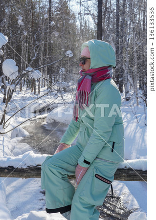 Woman in warm winter clothing sits outdoors in a snowy forest, wearing a hood, scarf, and sunglasses in bright cold sunlight 136133536