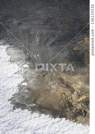 Snowy ice crystals meet clear flowing water, creating contrasting textures of white frost and warm-toned streambed beneath 136133538