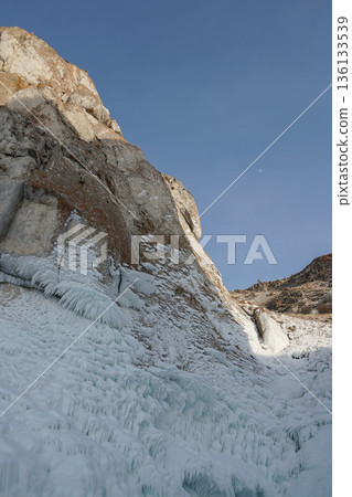 Frozen rock cliff covered with thick ice formations and icicles rises against a clear blue winter sky 136133539