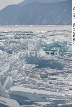 Sharp translucent ice plates, turquoise shards form dramatic frozen patterns on the surface of Lake Baikal with distant mountains in soft winter light 136133549