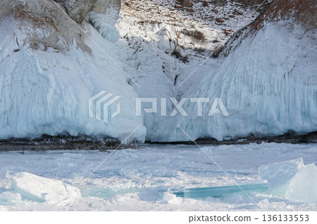 Frozen shoreline of Lake Baikal with massive ice-covered cliffs and layered turquoise ice plates in the foreground under cold daylight 136133553