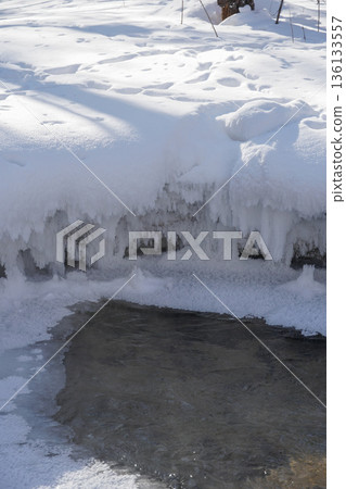 Frozen stream edge with thick snow layer and hanging ice formations above clear flowing water, soft winter light and calm mood 136133557