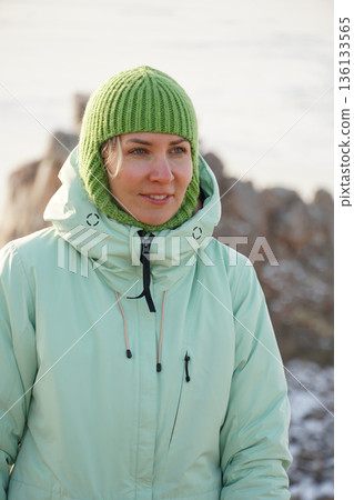 Smiling woman in a green knit hat and warm winter jacket stands outdoors near Lake Baikal in soft cold light Smiling woman in a green knit hat and warm winter jacket stands outdoors near Lake Baikal in soft cold light 136133565