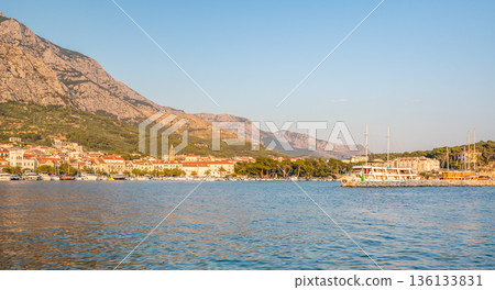 Panorama view of Makarska city, Croatia during the sunset. View with small boats and Biokovo mountains in the background. Summer weather, soft warm colors. Panorama view of Makarska city, Croatia during the sunset. View with small boats and Biokovo mountains in the background. Summer weather, soft warm colors. 136133831