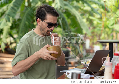 Freelancer drinking coconut water while working on laptop in tropical cafe Freelancer drinking coconut water while working on laptop in tropical cafe 136134576