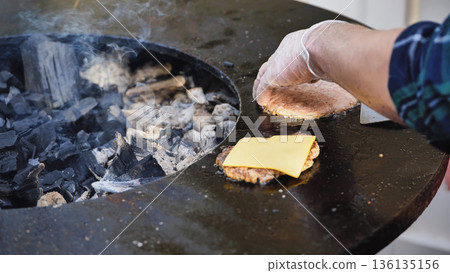 Person cooking beef burger patties on a charcoal bbq grill with melting cheese 136135156